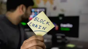 Bitunix A person holds a yellow sticky note with "BLOCK CHAIN" written on it in front of a computer screen, highlighting the concept of blockchain technology, with the background blurred.