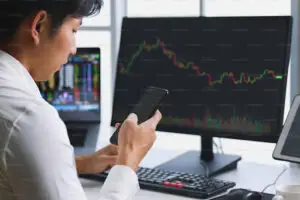 Bitunix A person sits at a desk looking at a smartphone, with two computer monitors displaying stock market charts, financial data, and crypto trading information in the background.