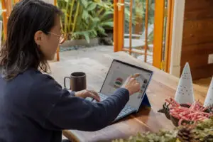 Bitunix A person sits at a wooden table using a stylus on a tablet with a keyboard, browsing a shopping website that supports smart contract wallets. A mug, candy canes, and holiday decorations are on the table; glass doors open to a garden outside.