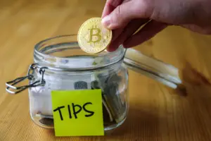 Bitunix A hand places a gold Bitcoin token into a glass jar labeled TIPS with a yellow sticky note. The jar, often used by those exploring crypto trading, also contains cash and coins and sits on a wooden surface. | Bitunix Bitunix A hand places a gold Bitcoin token into a glass jar labeled TIPS with a yellow sticky note. The jar, often used by those exploring crypto trading, also contains cash and coins and sits on a wooden surface.