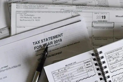 Bitunix A pen rests on a 2019 tax statement surrounded by paperwork, including 1099-MISC forms, a planner with a handwritten mileage log and business expenses, and notes about Crypto Taxes.