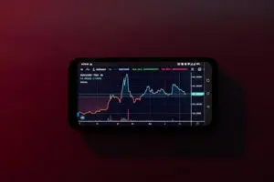 Bitunix A smartphone displays a financial chart with two fluctuating lines, one red and one blue, hinting at cryptocurrency trends or staking performance. The device rests on a maroon surface against a dark background.