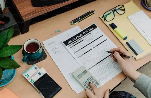 Bitunix A person fills out a tax application form at a desk with a calculator, pen, documents, highlighters, glasses, euro banknotes in a wallet, a cup of tea, and a potted plant—carefully reviewing details related to crypto taxes.