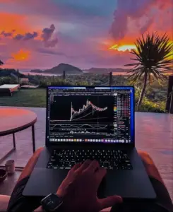 Bitunix A person uses a laptop displaying spot prices and futures prices on a candlestick stock chart, sitting outdoors at sunset with a scenic view of mountains, palm trees, and a vibrant, colorful sky. A cup and table are nearby.