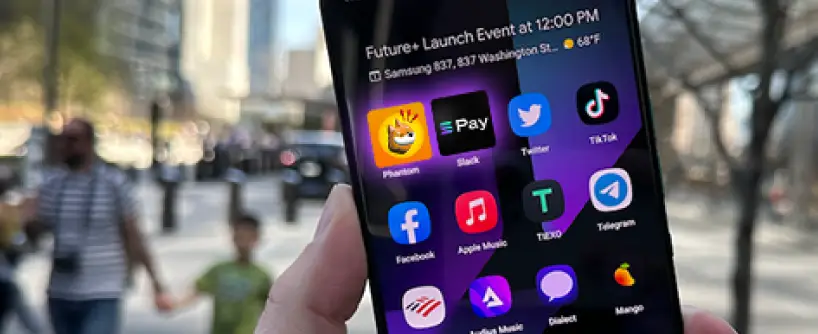 Bitunix A hand holds a smartphone displaying various app icons, including Solana, and a notification about a Future+ Launch Event at 12:00 PM. A city street with people walking in the background is slightly blurred.