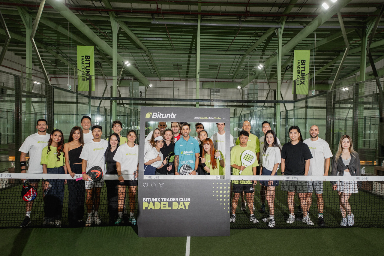 A group of people stand on an indoor padel court, posing behind a sign that reads BITUNIX TRADER CLUB PADEL DAY. Most are wearing sportswear, and some hold padel rackets. Bright green Bitunix banners hang in the background.